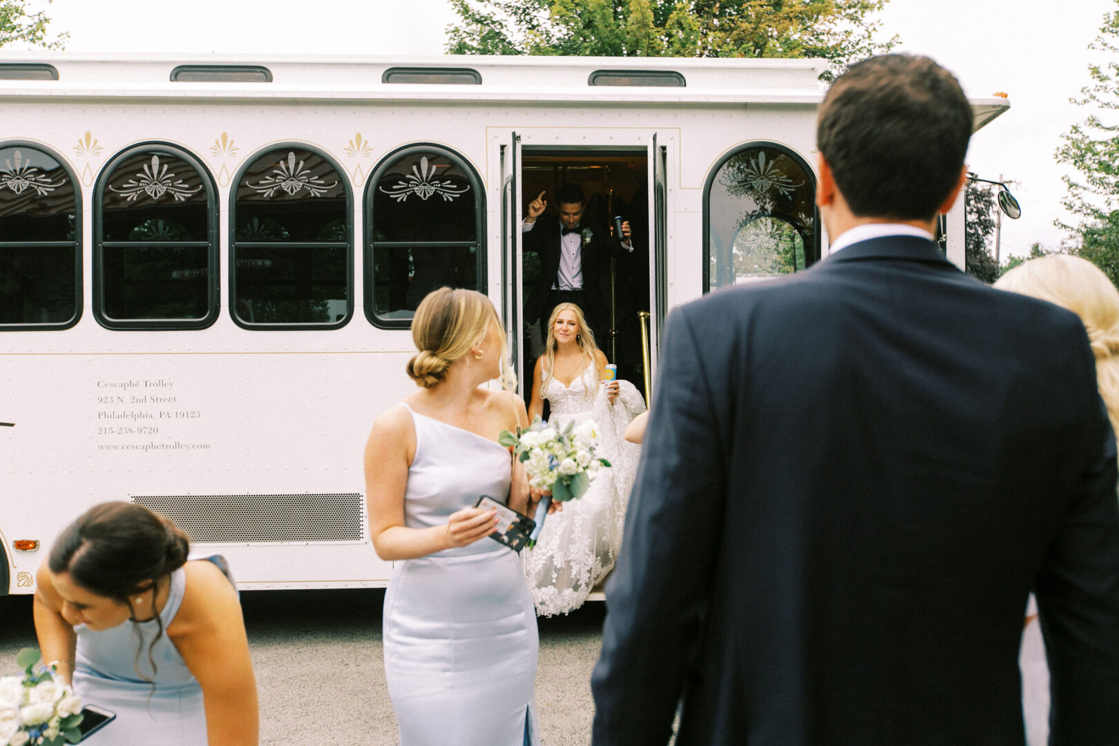 A bride emerges from a white trolley at Normandy Farm Wedding, followed by a groom flashing a peace sign. Three bridesmaids clutch bouquets nearby, surrounded by guests in formal attire.