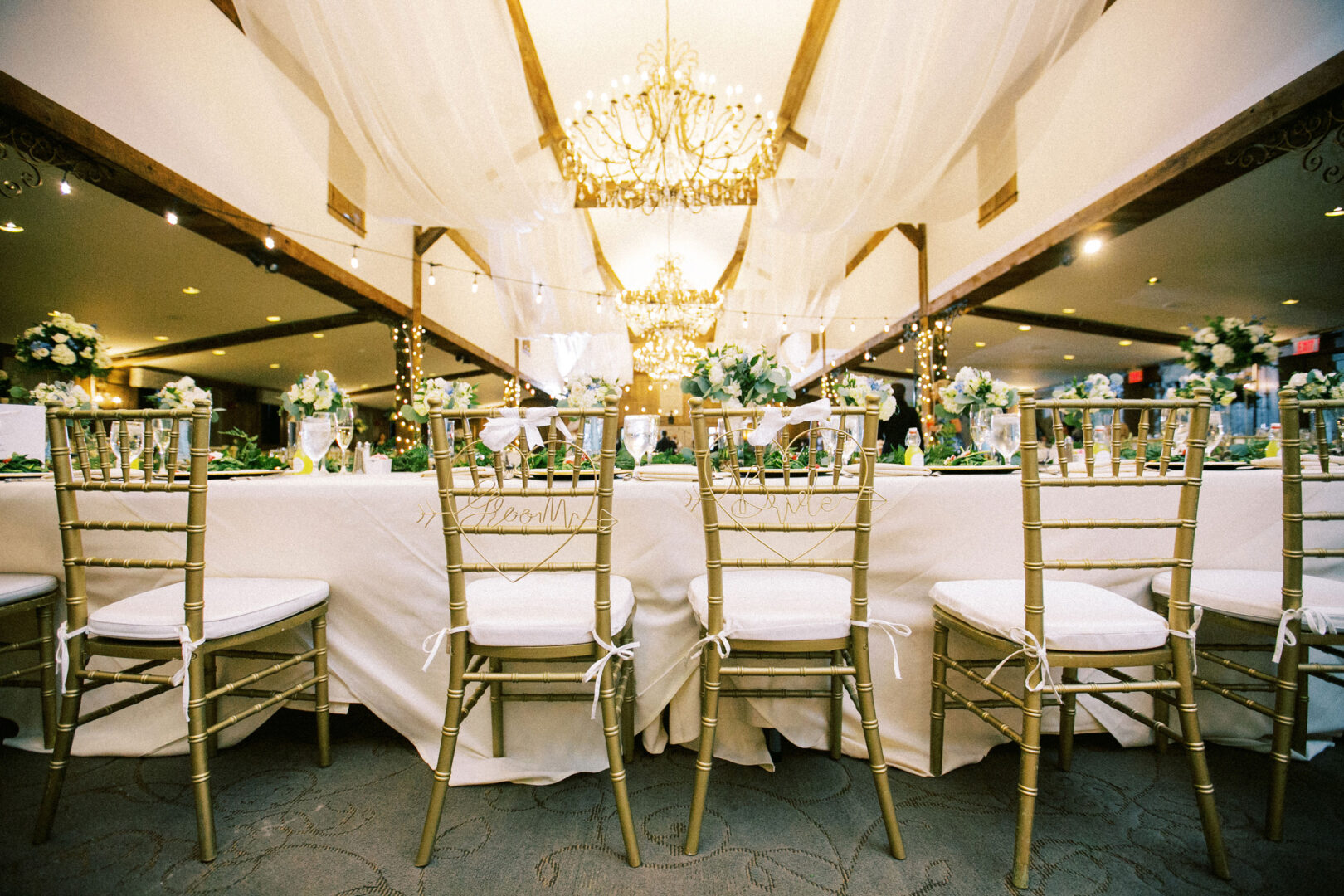 Experience the charm of a Normandy Farm Wedding with an elegant reception setup featuring a long dining table, gold chiavari chairs, stunning floral centerpieces, and a chandelier casting a warm glow overhead.