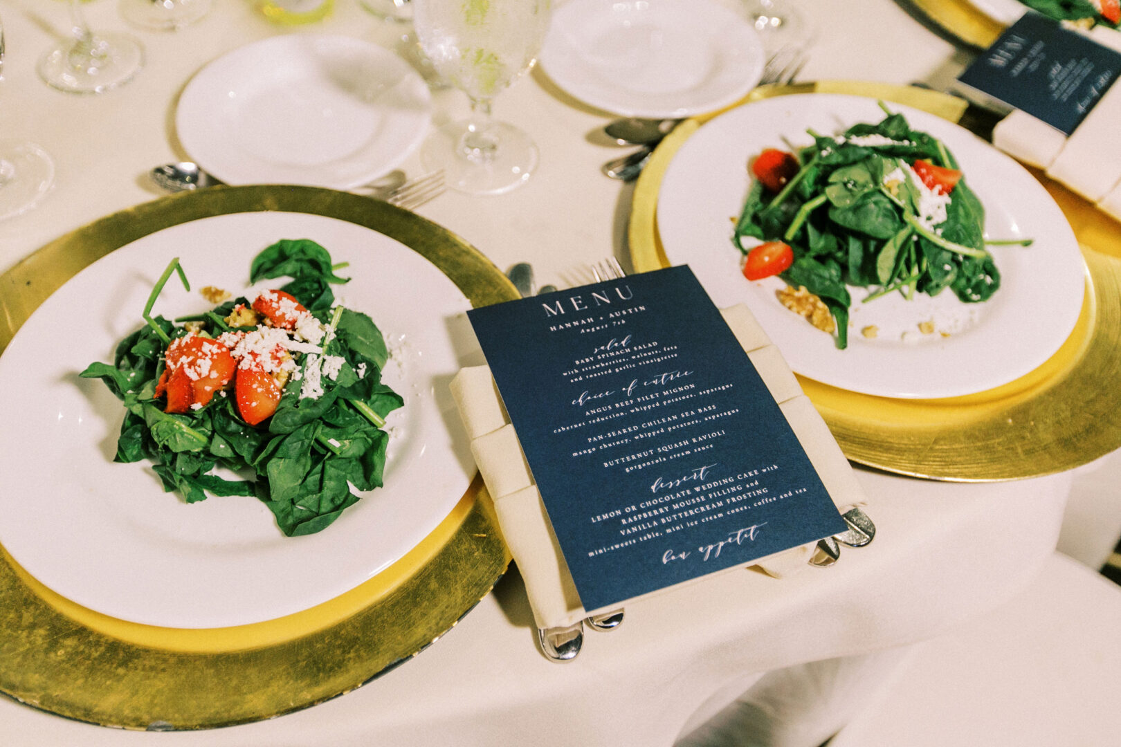 Two plates of salad with spinach, tomatoes, and cheese sit elegantly on a table at the Normandy Farm wedding. A menu is placed next to the plates on gold chargers.