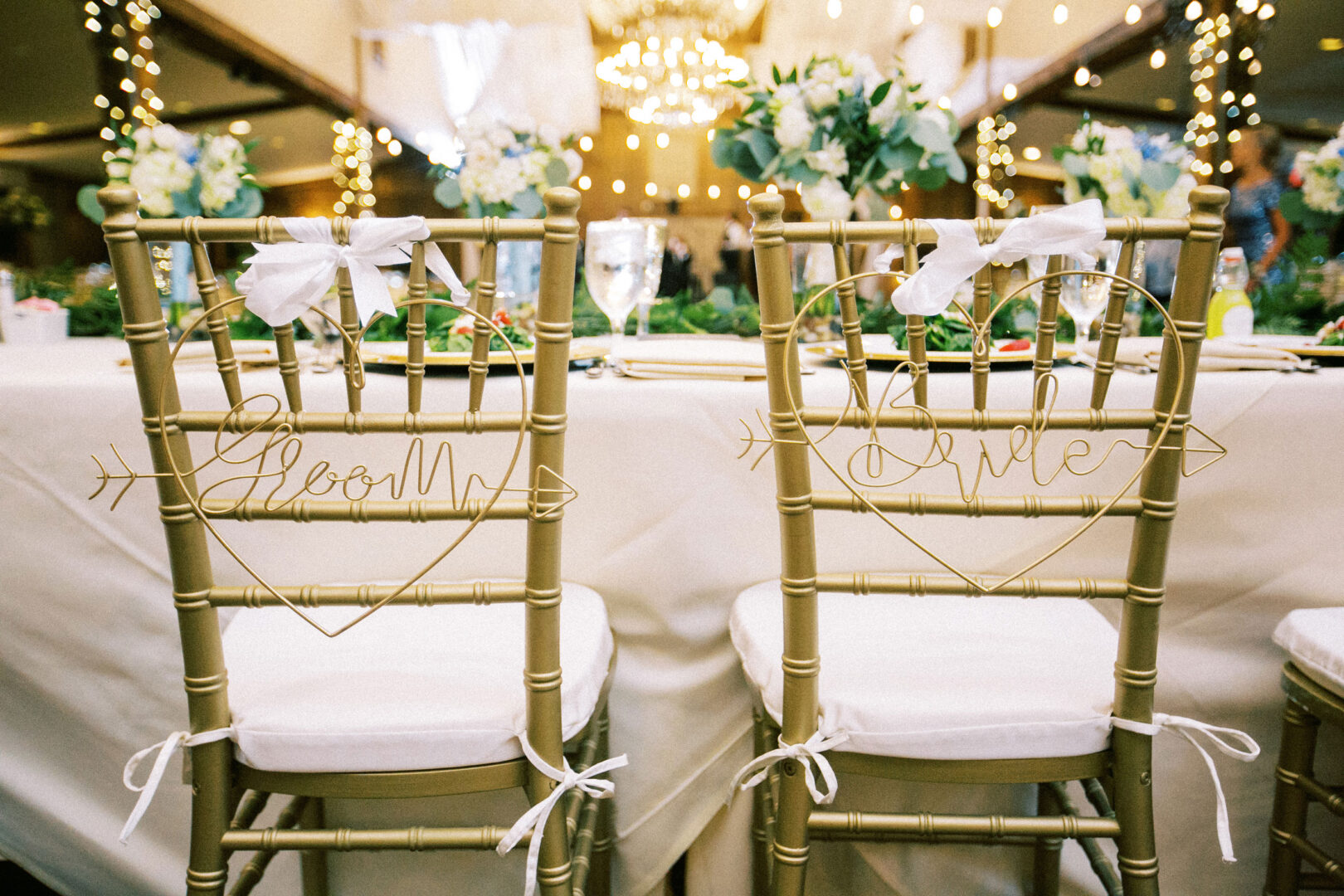 At a picturesque Normandy Farm wedding, two gold chairs with "Groom" and "Bride" signs are elegantly adorned with white ribbons, sitting before a table beautifully set with flowers and sparkling glassware.