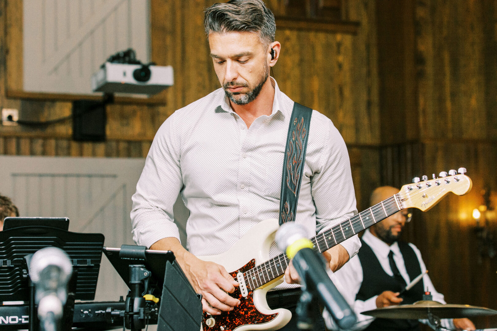 In a room exuding the charm of a Normandy Farm wedding, a man in a white shirt skillfully plays an electric guitar, with band equipment and a drummer providing the rhythmic backdrop.