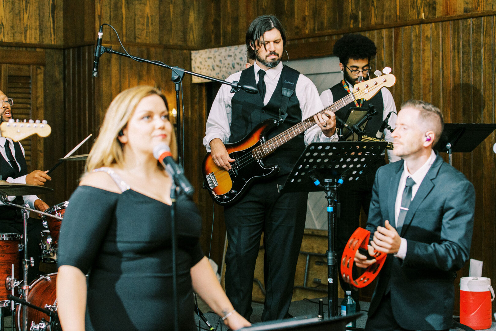 At a Normandy Farm wedding, a band performs indoors with a woman singing, a man strumming the bass guitar, another keeping rhythm with a tambourine, and musicians adding depth in the background.