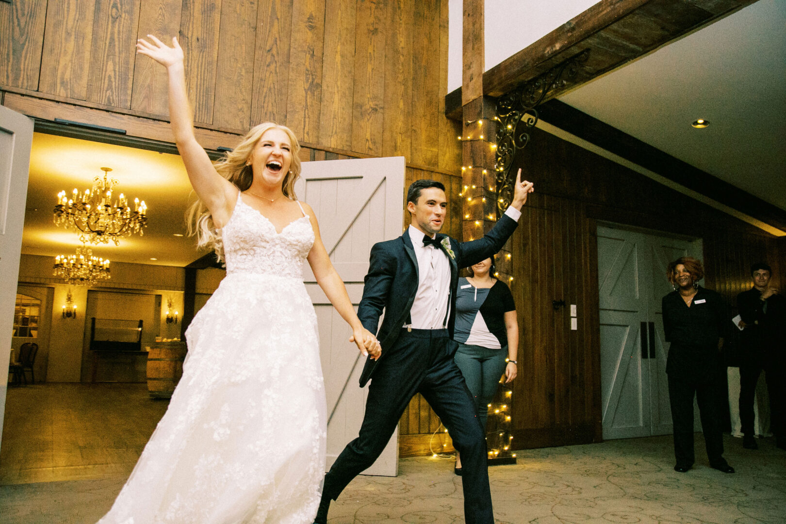 A bride and groom joyfully enter the Normandy Farm Wedding reception hall, holding hands. The bride raises her arm, and guests cheer them on. The room is adorned with chandeliers and string lights, setting a magical scene.