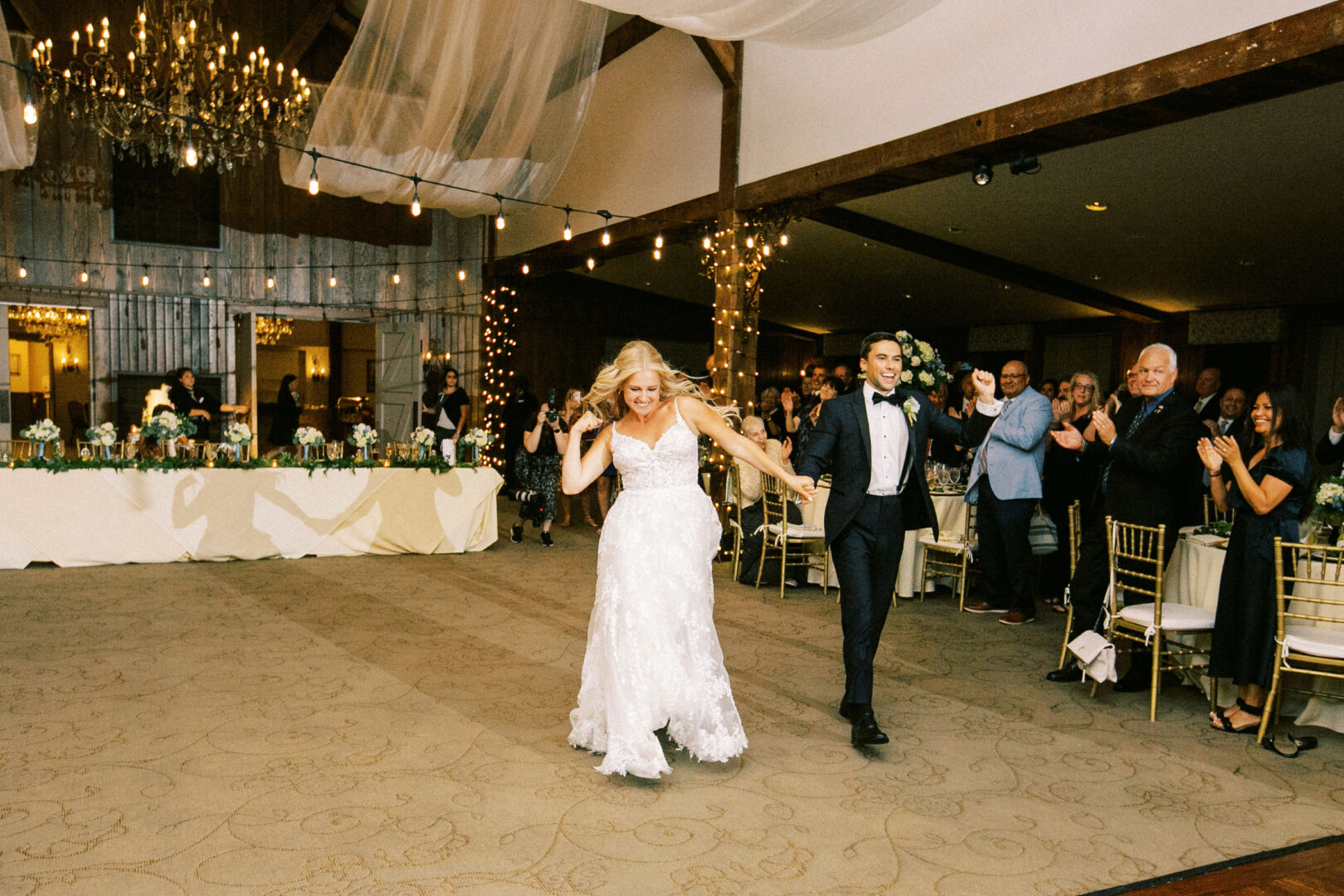 A bride and groom joyfully enter the beautifully adorned Normandy Farm Wedding reception hall, holding hands, as guests applaud.