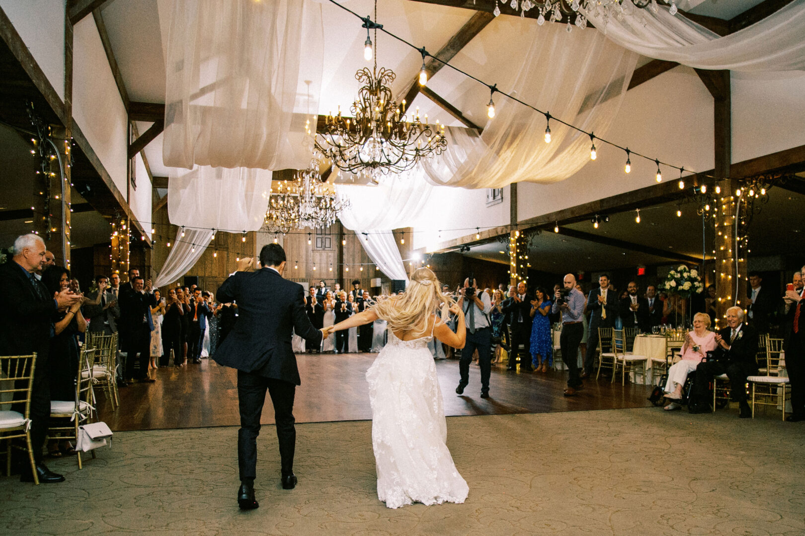 A bride and groom hold hands and walk into the beautifully decorated Normandy Farm Wedding reception hall, with guests applauding around them.