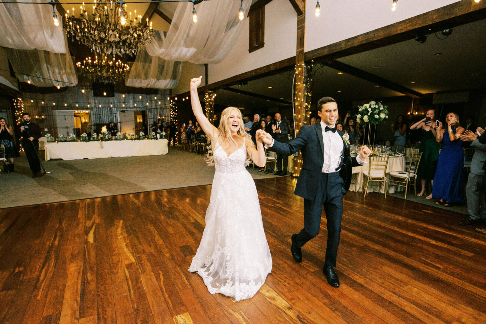 A bride and groom celebrate their Normandy Farm Wedding as they enter a reception hall filled with guests. They hold hands, and the bride raises her arm in excitement. The room features chandeliers and wooden flooring, creating an elegant atmosphere.