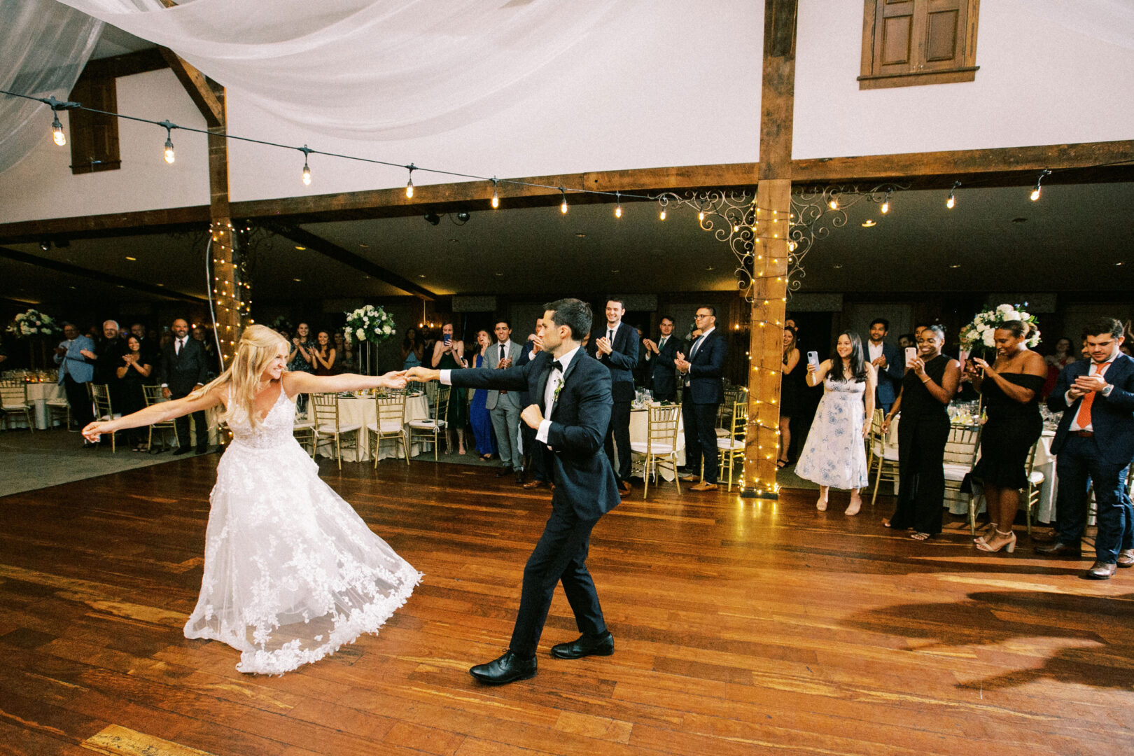 At their Normandy Farm wedding, the bride and groom dance energetically on a wooden floor, surrounded by applauding and smiling guests in the beautifully decorated venue.