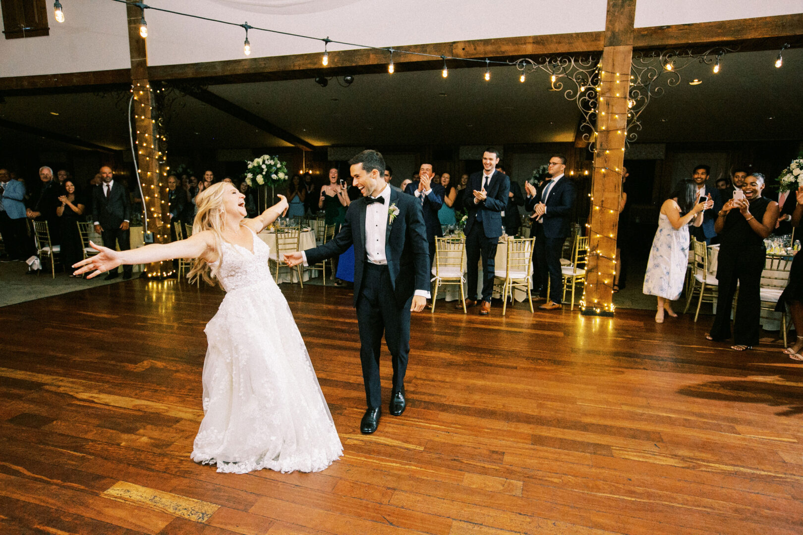 A couple in formal attire dances joyfully on the wooden floor of a Normandy Farm Wedding, surrounded by applauding guests and shimmering string lights.