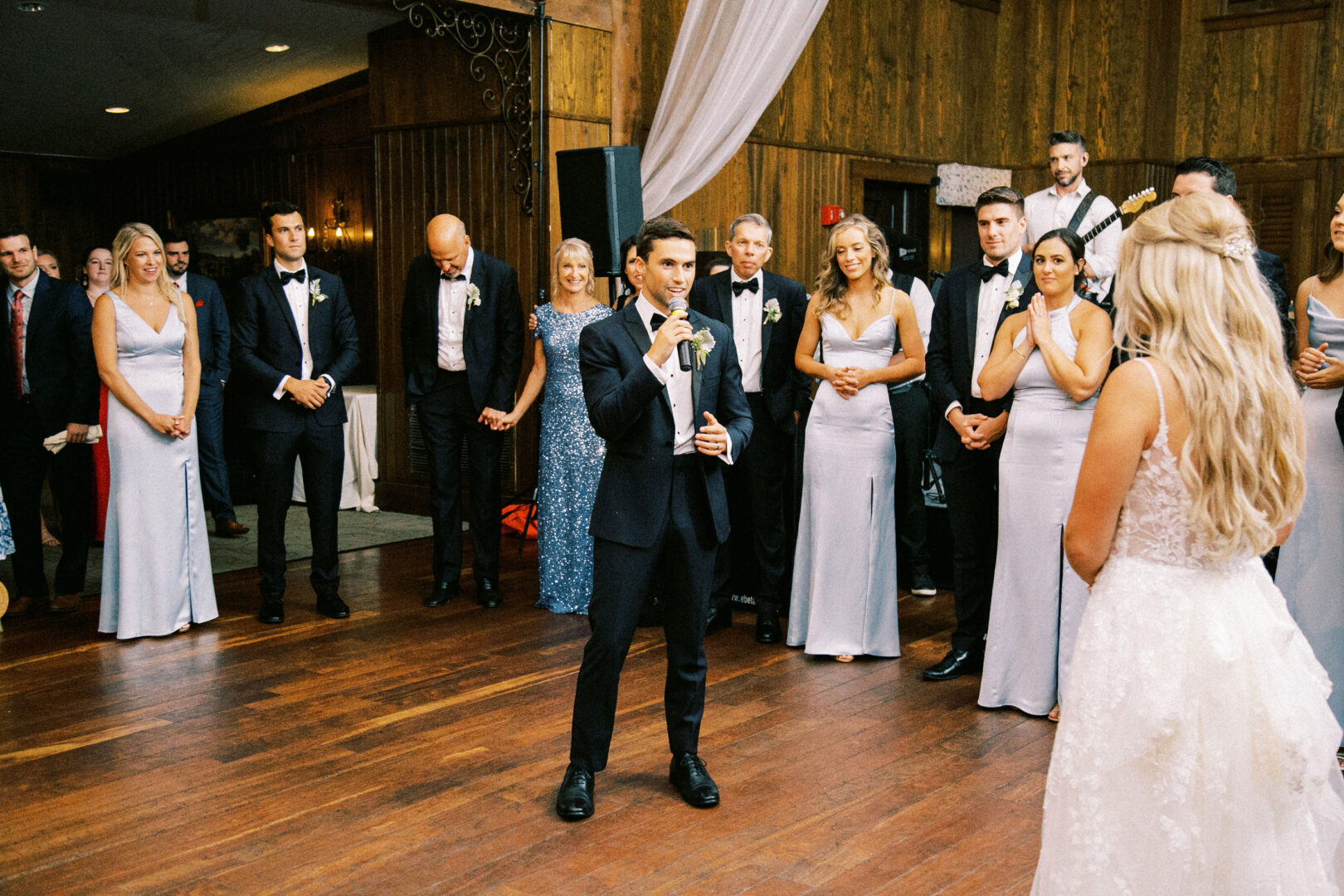 A man in a suit gives a speech with a microphone at the Normandy Farm wedding reception. The bride and other guests stand and listen attentively in the elegant, wood-paneled room.