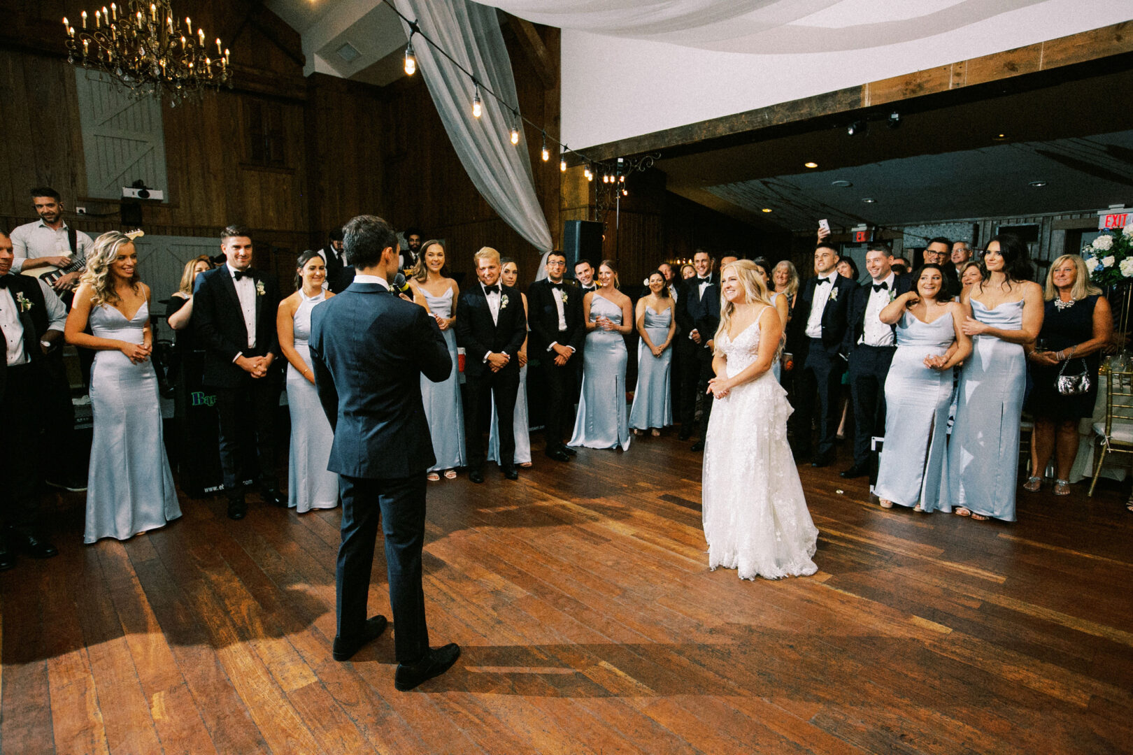 A bride and groom stand facing each other in the center of the Normandy Farm ballroom, surrounded by a crowd of guests in formal attire.