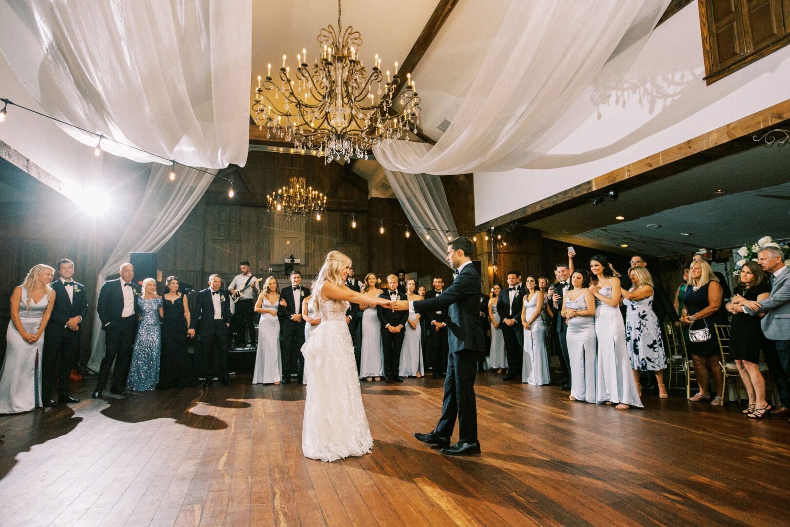 At their Normandy Farm wedding, the bride and groom dance gracefully under chandeliers in a beautifully decorated hall, surrounded by guests who watch with joy.