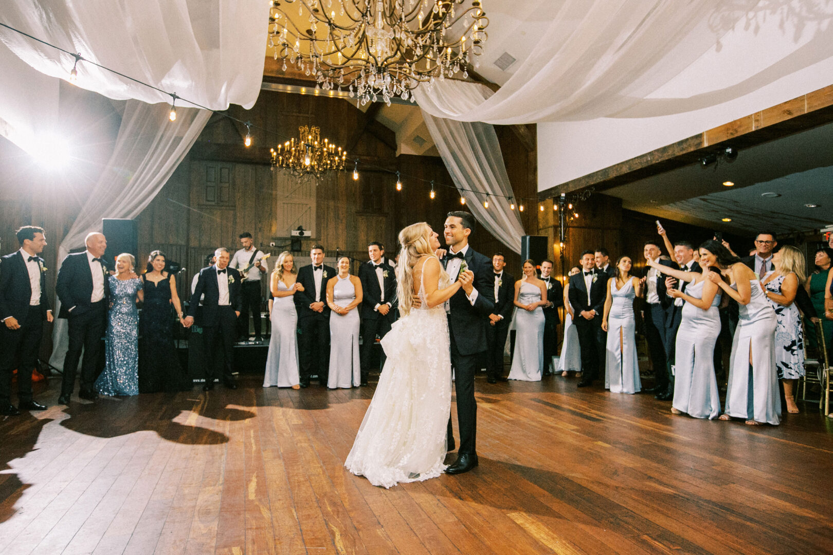 At their Normandy Farm wedding, the bride and groom share a magical first dance in a large hall, surrounded by their wedding party and guests, beneath shimmering chandeliers and elegantly draped fabric.