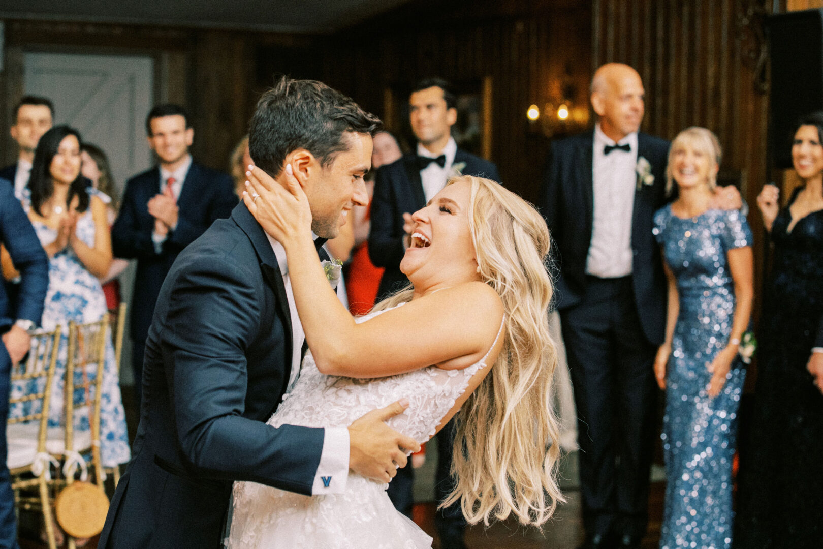 At a delightful Normandy Farm wedding, the bride and groom dance joyfully in the center of a crowded room, surrounded by smiling guests in formal attire.