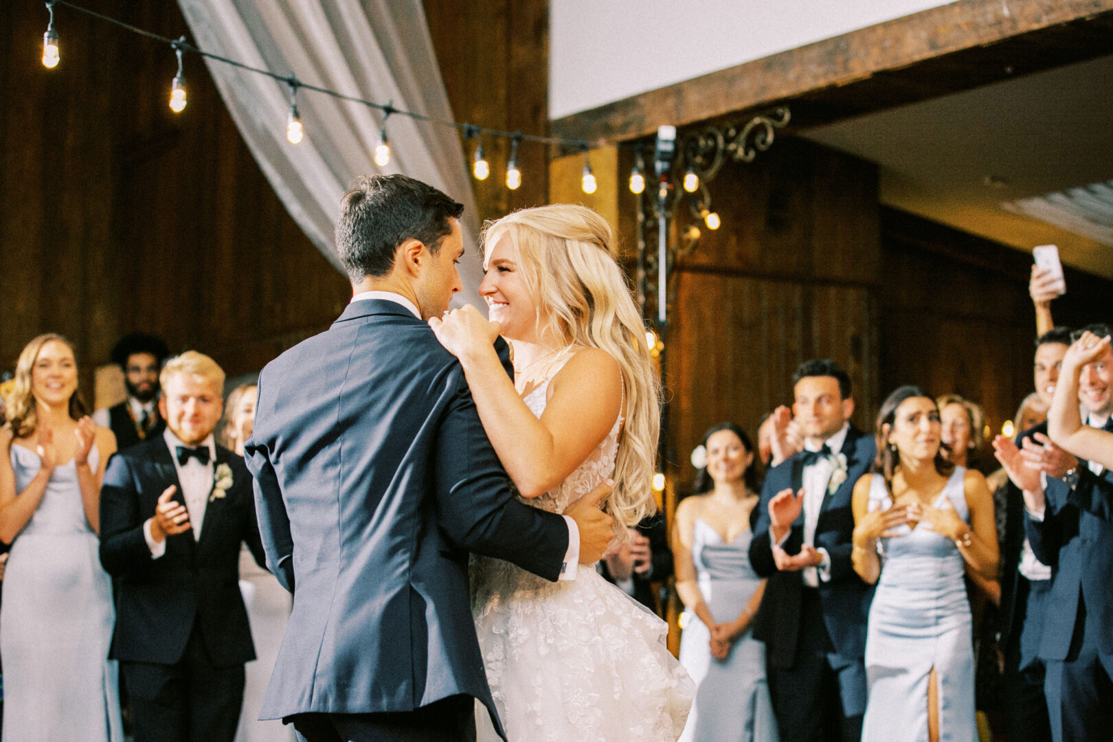 At a Normandy Farm wedding, the bride and groom dance surrounded by smiling guests in formal attire, inside a warmly lit venue.