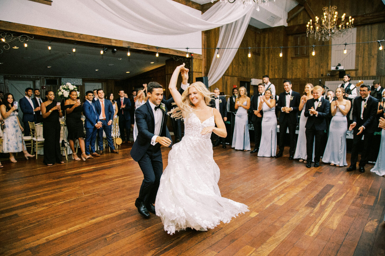 At a Normandy Farm wedding, a couple in elegant attire sways gracefully at the center of the reception hall, surrounded by applauding guests.