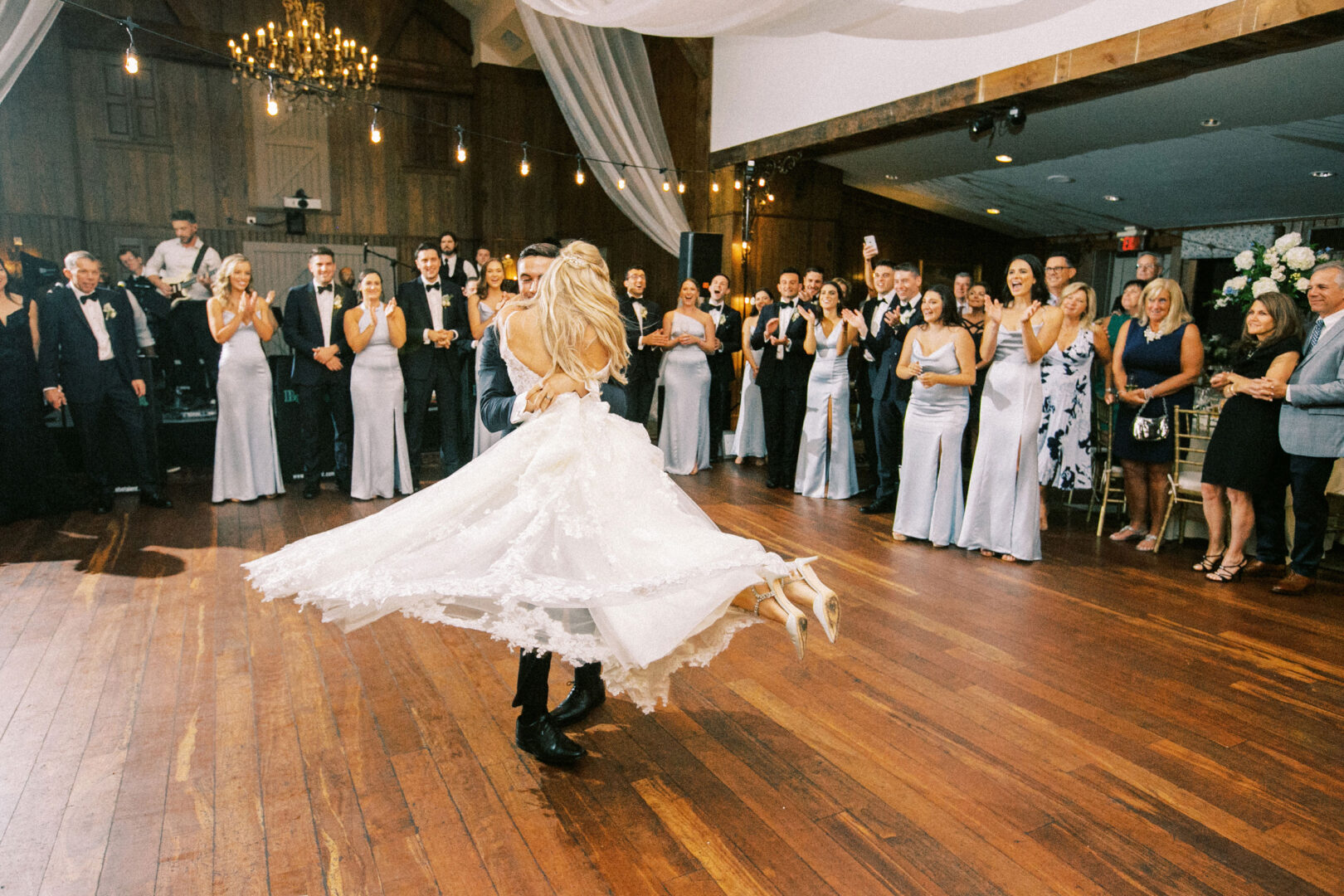 At a charming Normandy Farm wedding, a bride and groom dance joyfully in the center of a wooden-floor ballroom, surrounded by smiling guests in formal attire.