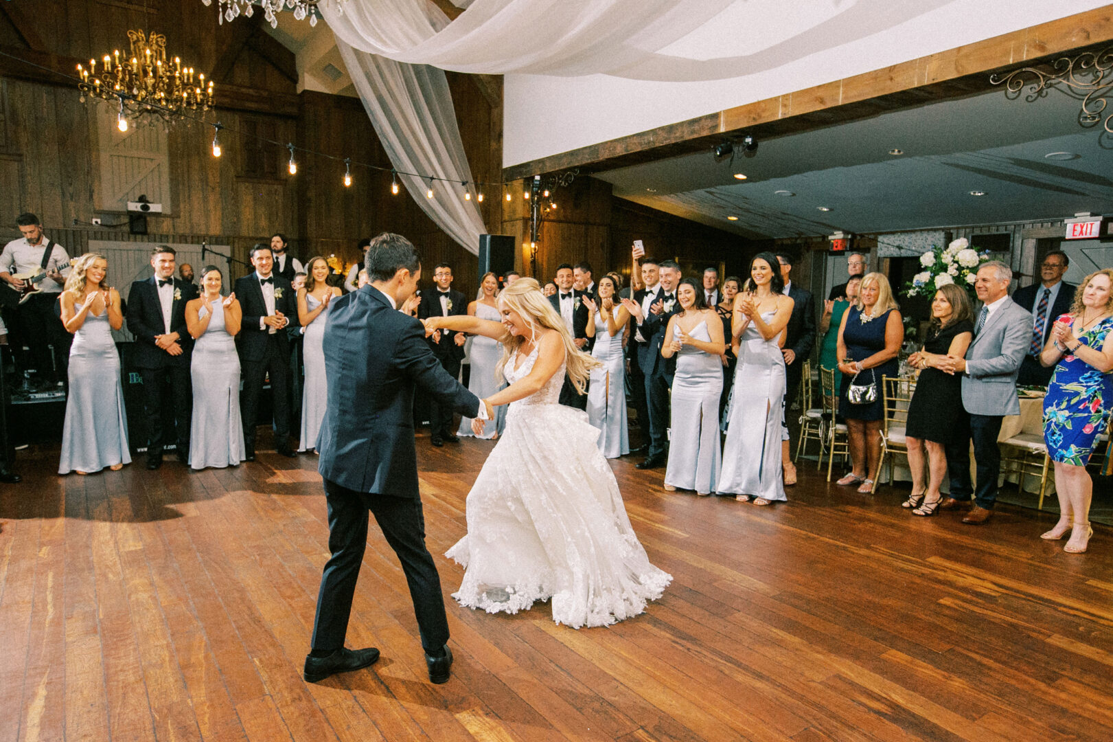 At a charming Normandy Farm Wedding, a bride and groom dance elegantly in a spacious room, surrounded by guests clapping and watching. The ceiling is beautifully adorned with drapes and chandeliers, adding to the warm and romantic atmosphere.