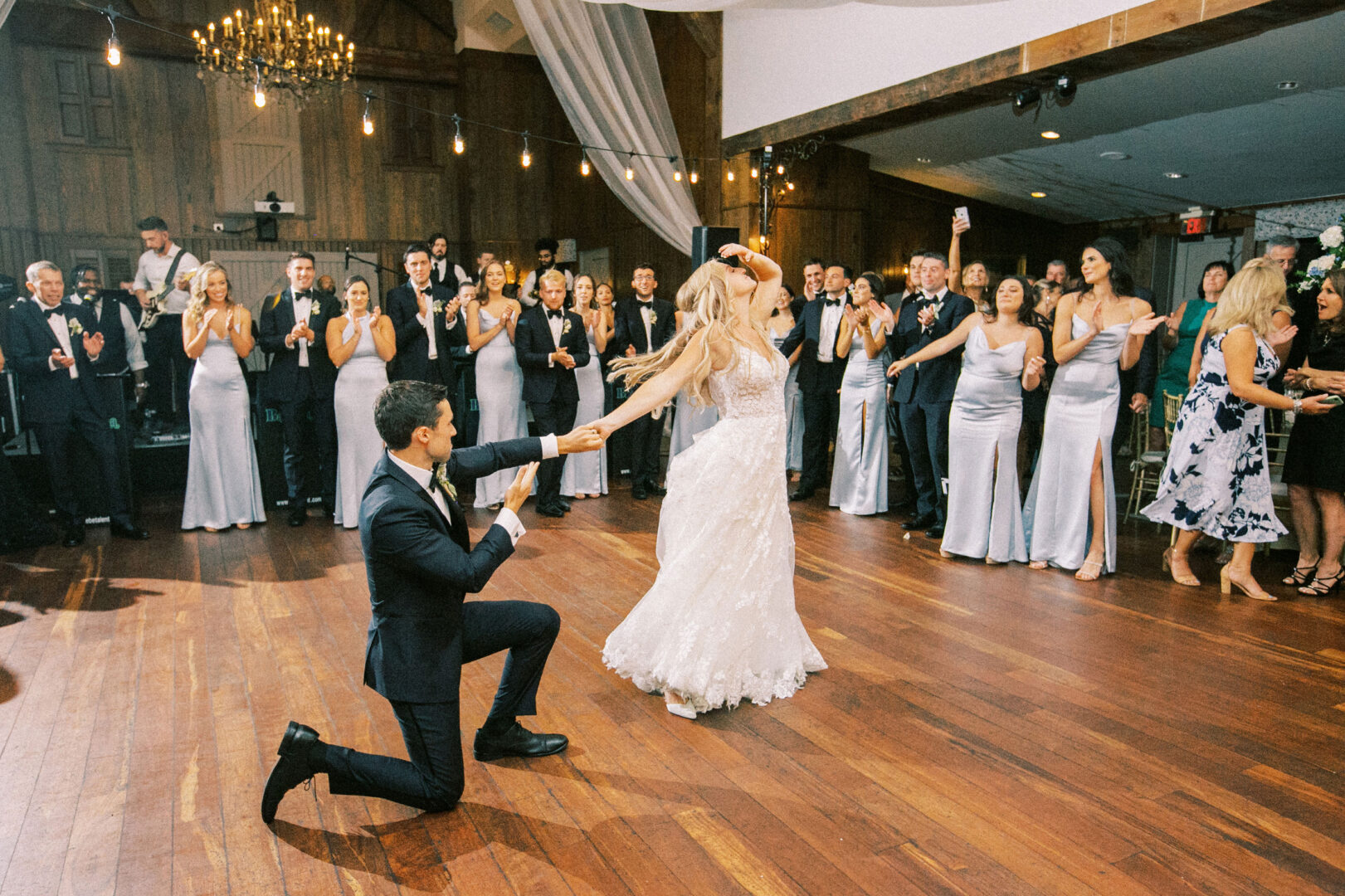 At a Normandy Farm wedding, the bride and groom twirl gracefully on the wooden floor, enveloped by guests in elegant formal attire who burst into enthusiastic applause.