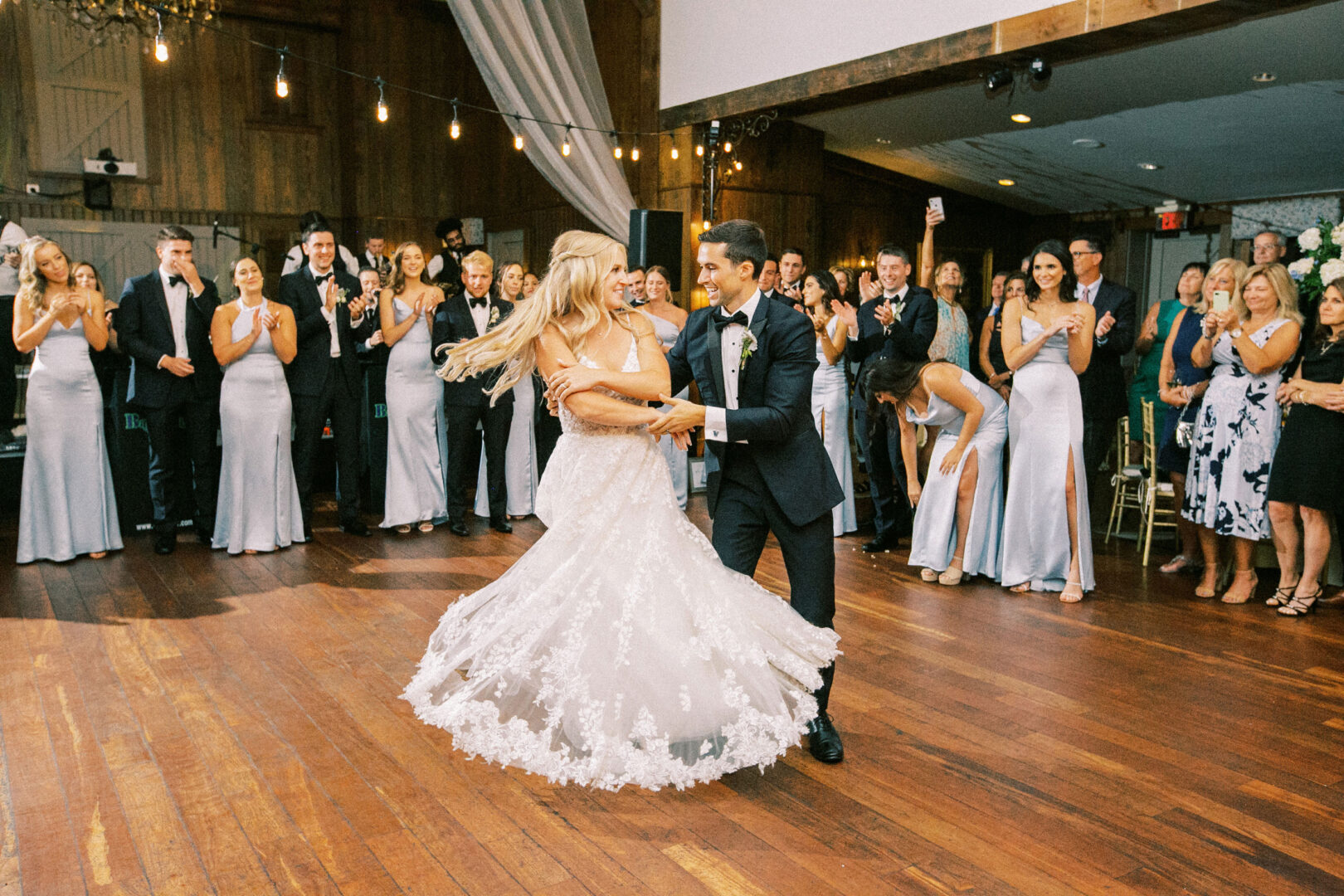 At their Normandy Farm wedding, a bride and groom dance in the center of a wooden floor, surrounded by applauding guests in formal attire.