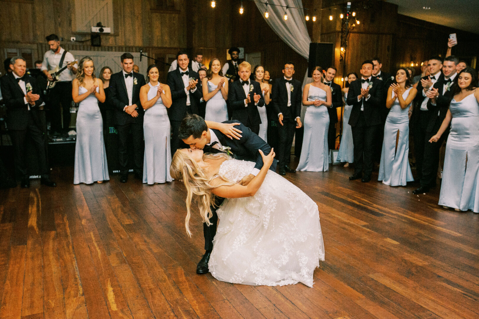 At a charming Normandy Farm wedding, a couple in elegant wedding attire dances gracefully, with the groom dipping the bride. Bridesmaids and groomsmen in formal wear stand clapping joyously in the background.