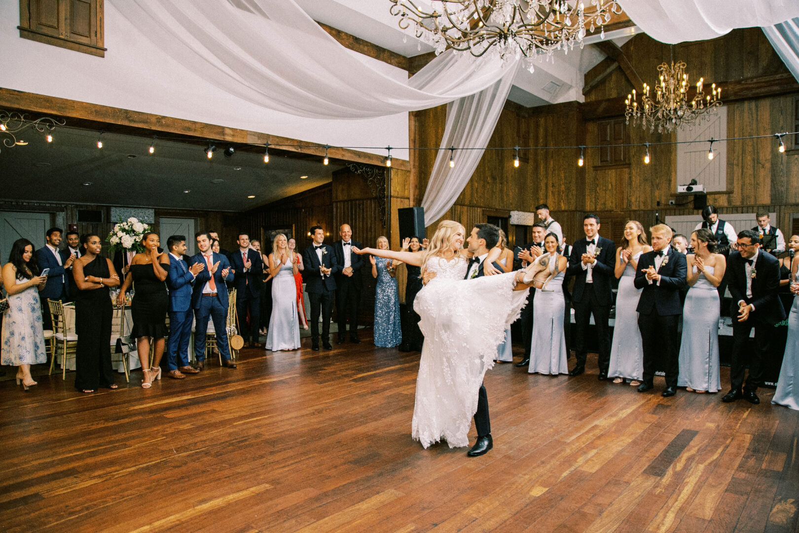 A couple performs a dance in the center of a room, surrounded by an audience applauding, at a Normandy Farm wedding venue with wooden floors and draped ceilings.