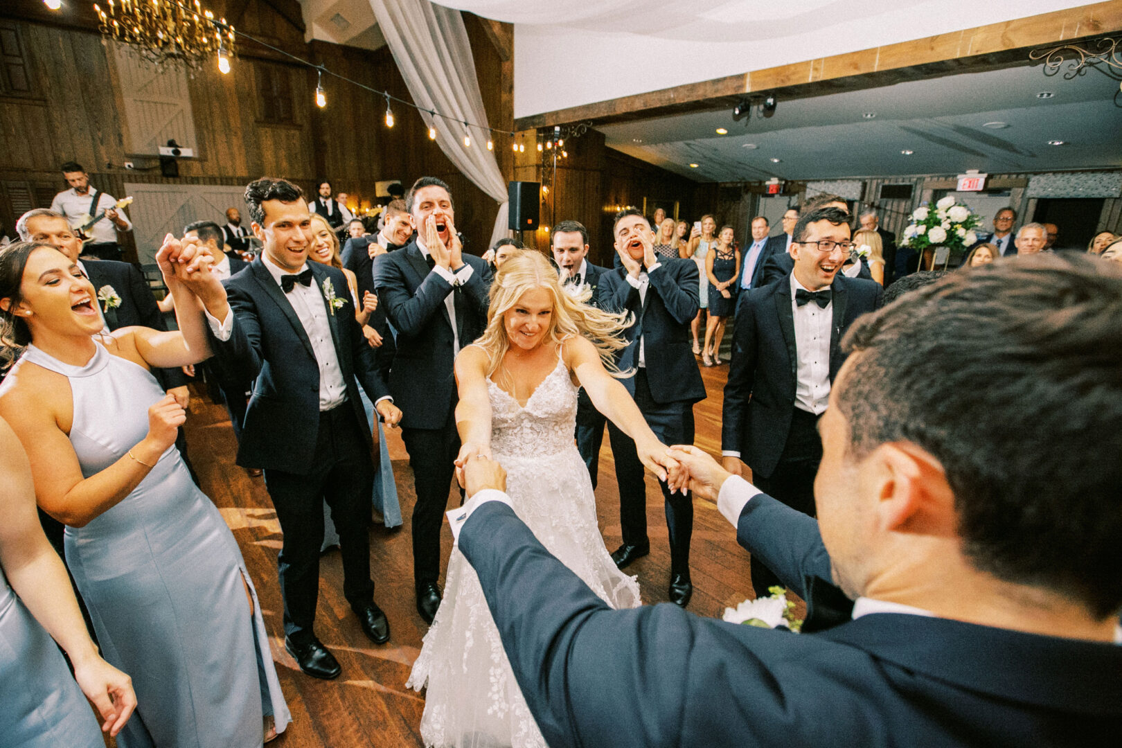 At their Normandy Farm wedding, the bride and groom dance enthusiastically, surrounded by cheering and clapping guests in elegant formal attire.