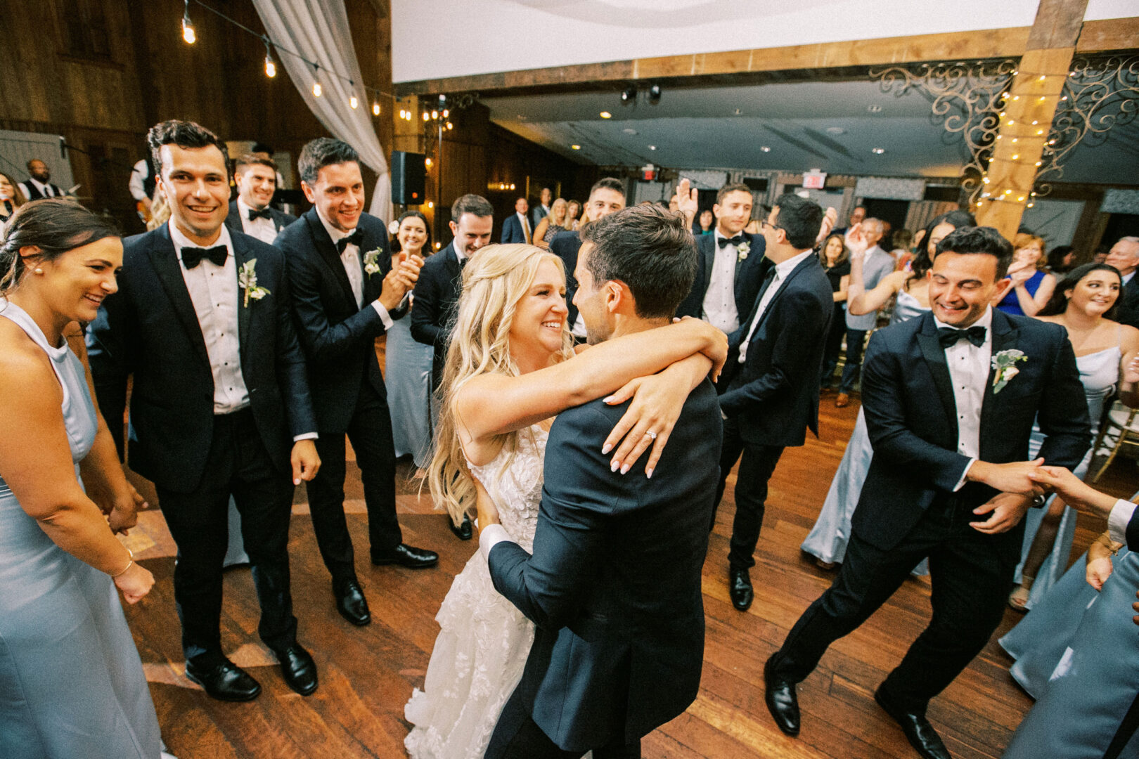 A bride and groom embrace in a lively wedding dance at their Normandy Farm Wedding, surrounded by smiling guests in formal attire on a wooden dance floor.