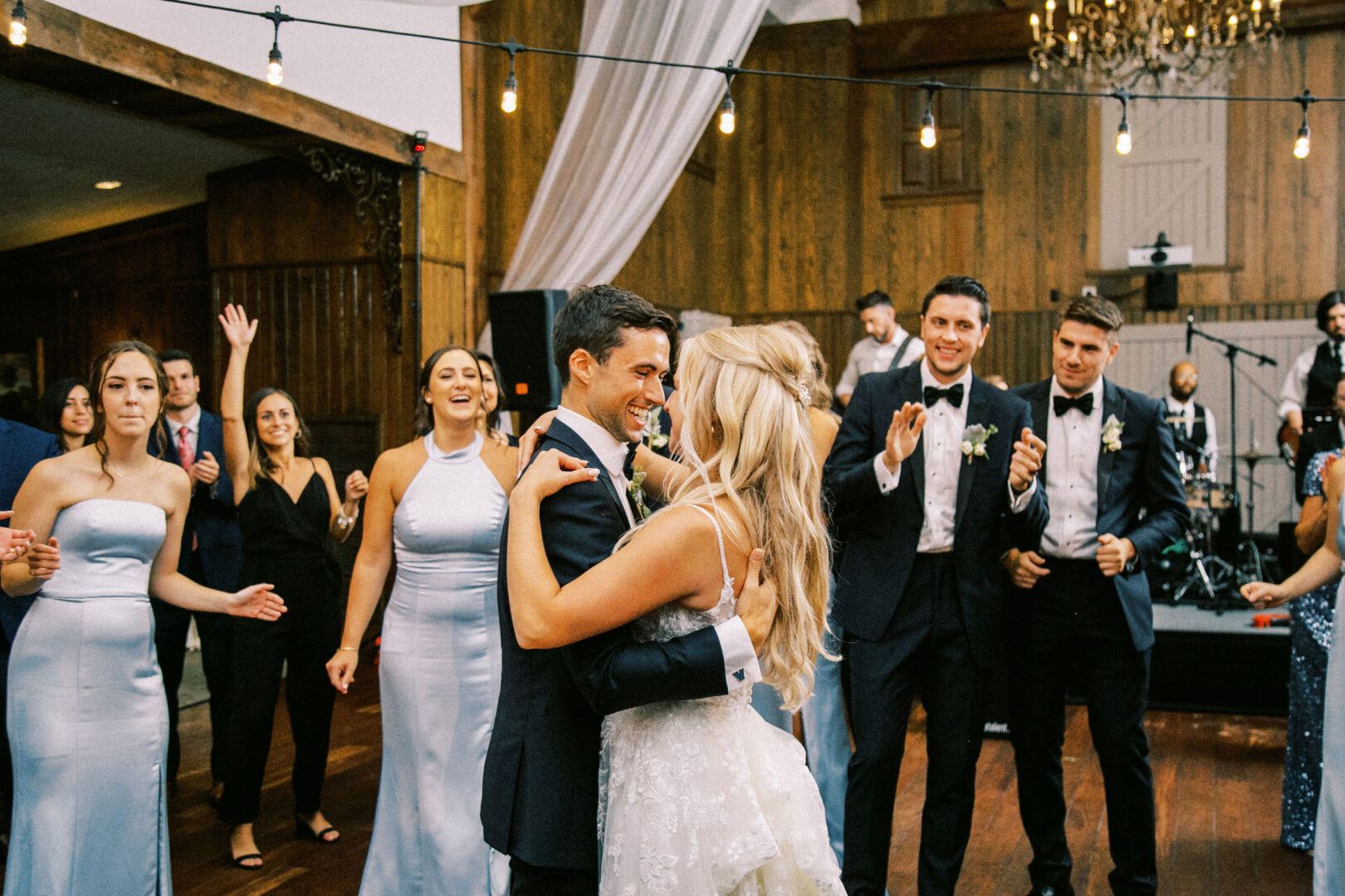 At a Normandy Farm Wedding, a couple dances at their reception, surrounded by smiling guests in formal attire under twinkling string lights.