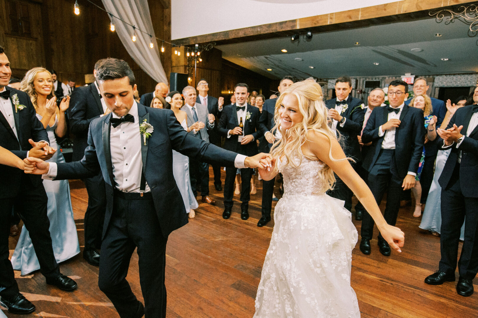 A bride and groom in formal attire dance joyfully on the wooden floor at their Normandy Farm wedding, surrounded by applauding guests in an elegant indoor venue.