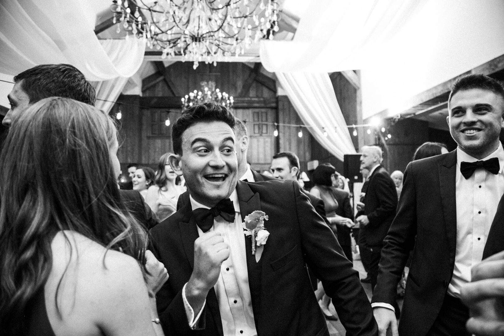 At the Normandy Farm Wedding, a group of people in formal attire danced and socialized beneath elegant chandeliers in the beautifully decorated indoor venue.