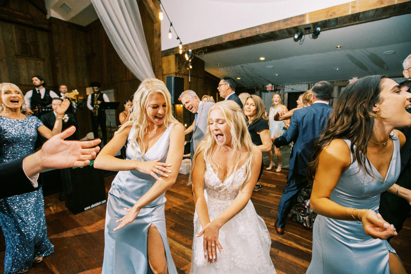 At a lively Normandy Farm wedding reception, the bride in a white dress dances energetically with her bridesmaids in blue dresses amidst the bustling indoor crowd.