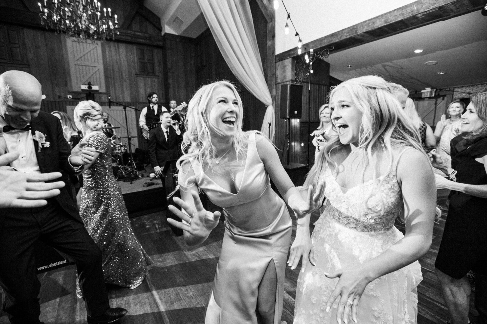 Two women in elegant dresses laugh and dance at a lively indoor event, reminiscent of a Normandy Farm wedding, with people in the background enjoying the celebration.