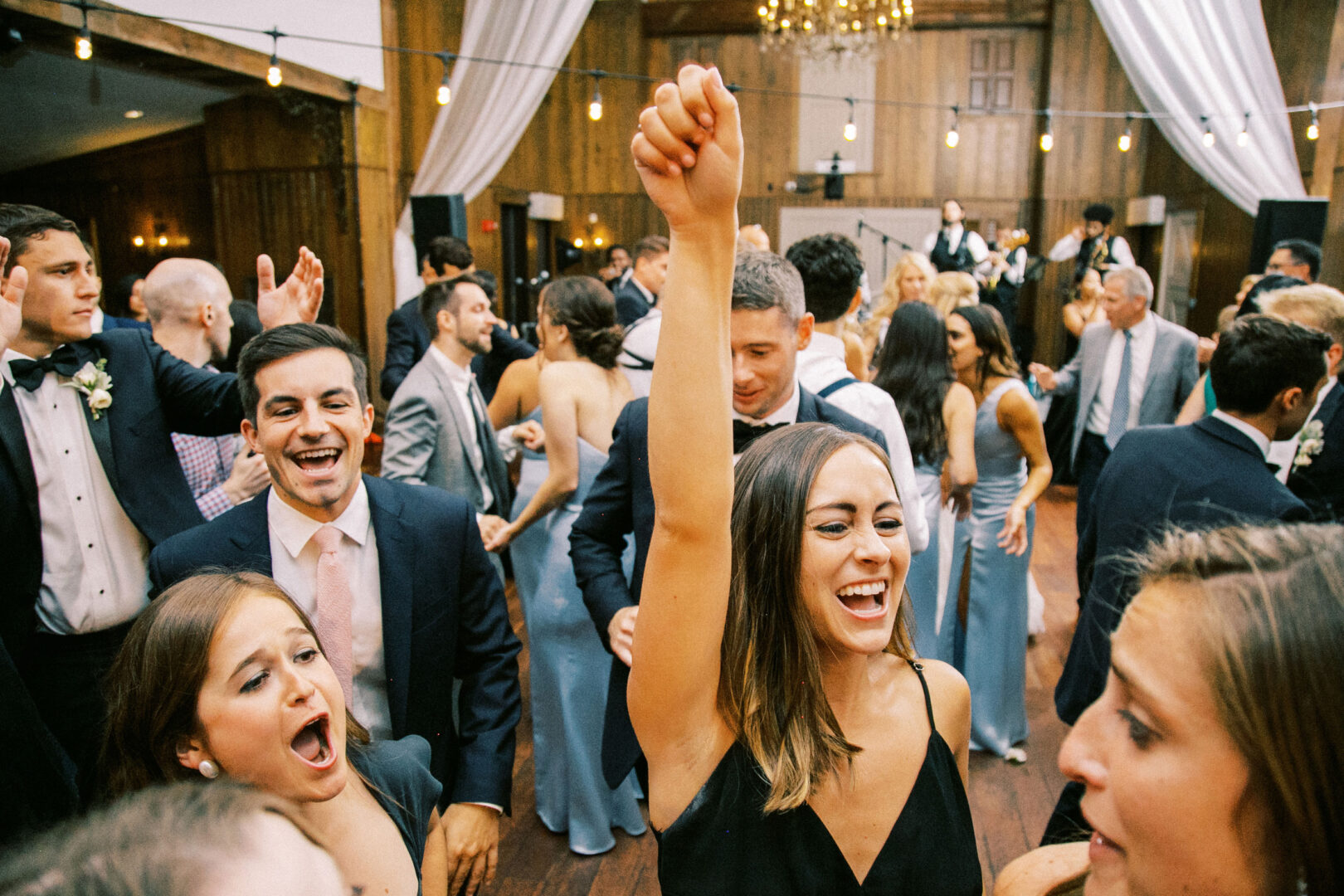 Guests in formal attire dance energetically at a Normandy Farm Wedding reception, with vibrant lights and skilled musicians creating an unforgettable atmosphere in the background.