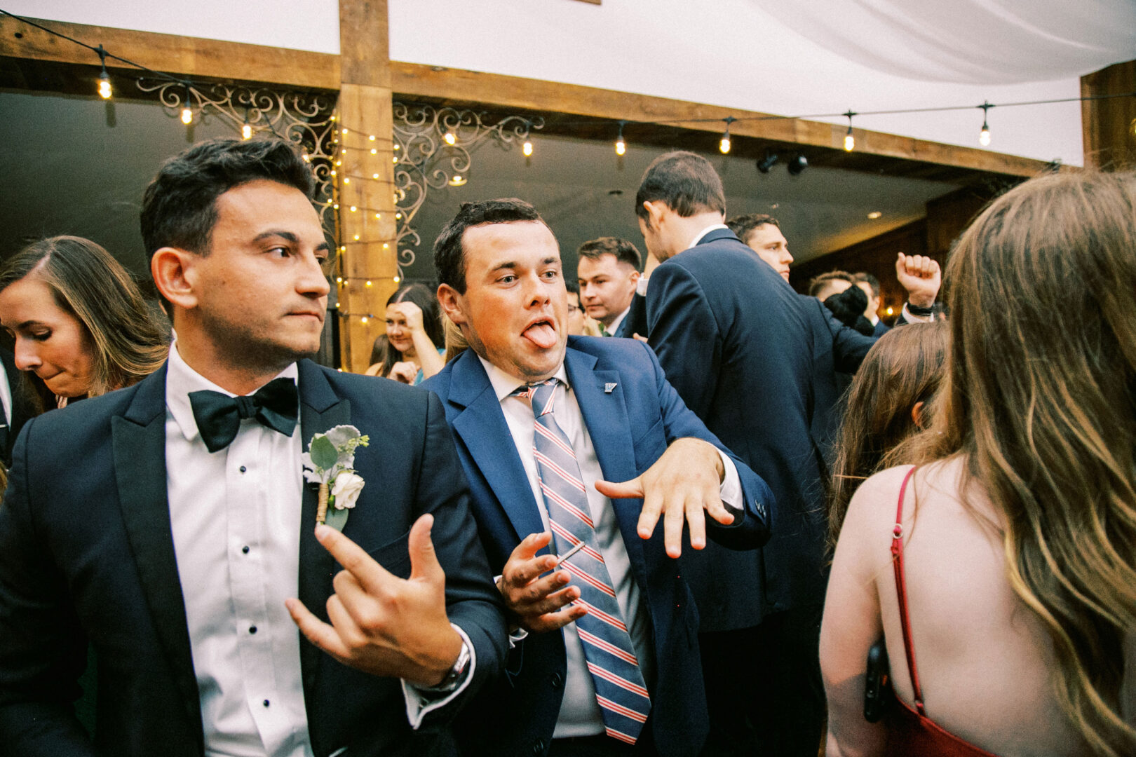 At a lively Normandy Farm wedding, a group of people in formal attire is energetically dancing indoors. The spotlight is on two men in suits making playful gestures, adding to the joyous atmosphere.