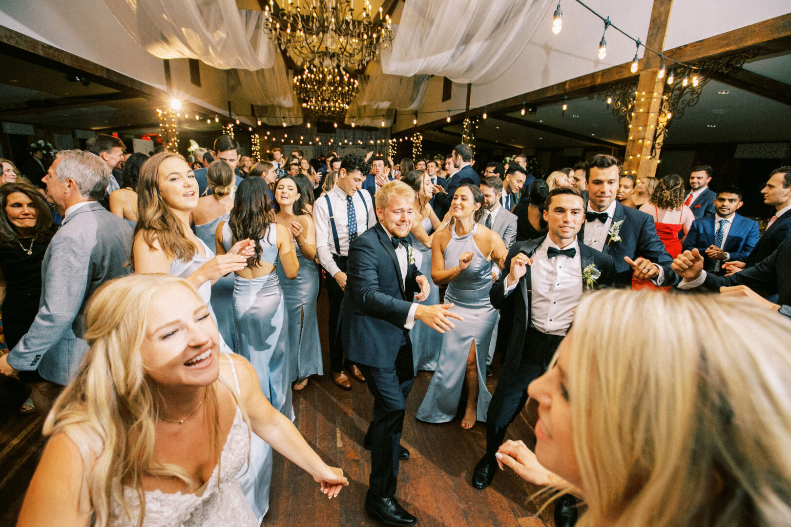 A large group of people, dressed in formal attire, are dancing and celebrating in a beautifully decorated ballroom at a Normandy Farm wedding, adorned with string lights and draped fabric.