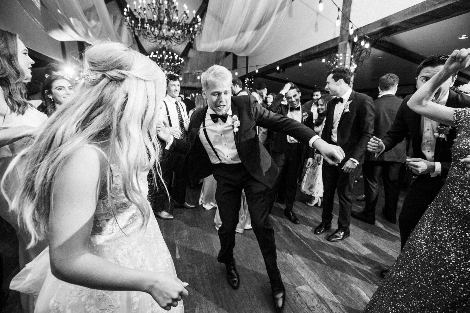 A group of people in formal attire revels in a lively dance at a celebration. Captured in black and white, the scene—reminiscent of a charming Normandy Farm wedding—features a chandelier and wooden beams above, adding to the timeless elegance.