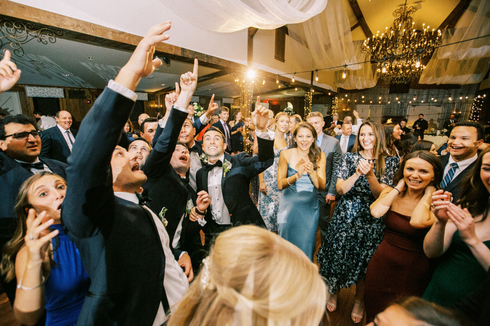 A joyful crowd of people, dressed in formal attire, celebrate together indoors under chandeliers and draped fabric at a Normandy Farm wedding. Some are cheering and pointing upwards.