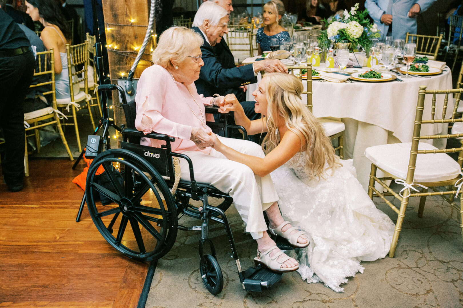 At a charming Normandy Farm wedding, a bride in a white dress kneels beside an elderly woman in a wheelchair, sharing a tender moment. Behind them, guests are seated at elegantly arranged tables, celebrating the union amid the rustic beauty of the venue.