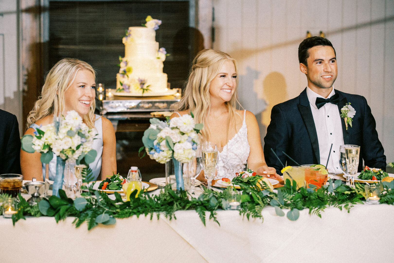 Three people in formal attire sit at a beautifully decorated table with food and drinks, evoking the charm of a Normandy Farm wedding. A cake, reminiscent of countryside elegance, is visible in the background.