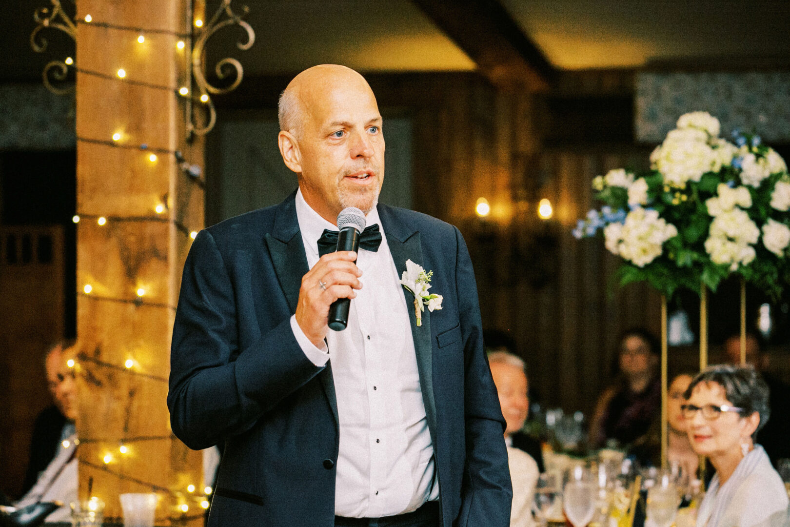 At a Normandy Farm wedding, a man in a suit speaks into a microphone at the event, with guests seated at elegantly arranged tables in the background.