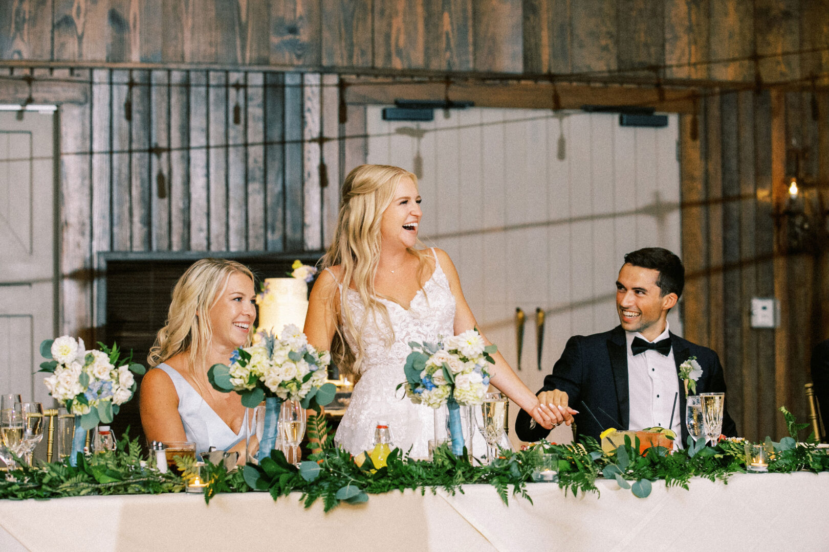 At a beautifully adorned Normandy Farm wedding table, the bride and groom sit with another guest. The bride is smiling, her hands firmly clasped with the groom's, as they savor this cherished moment together.