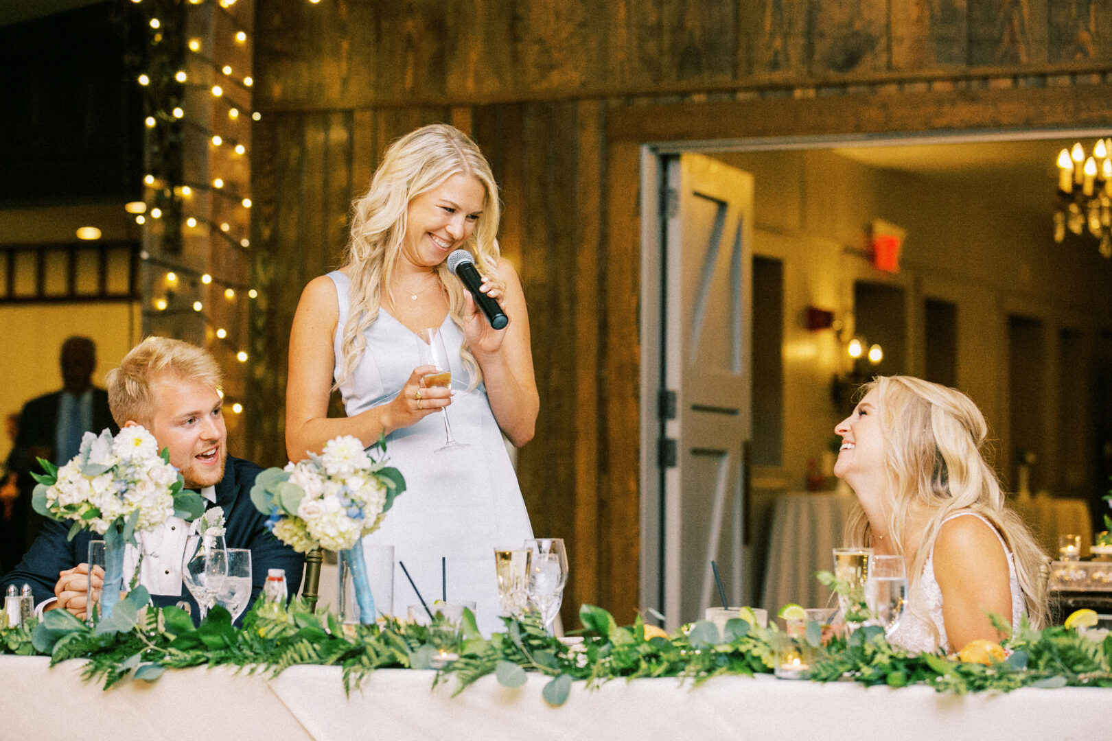 At the charming Normandy Farm wedding, a woman in a white dress stands gracefully and speaks into a microphone. Nearby, a man and a woman sit at their flower-adorned table, listening and smiling amid the lush greenery.