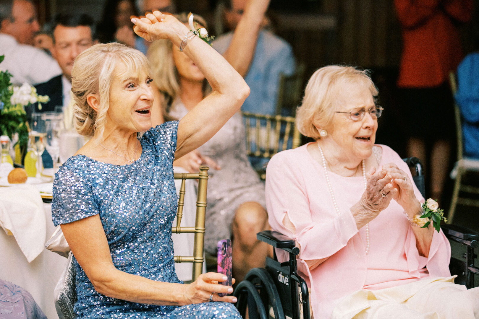 At a Normandy Farm wedding, two women radiate joy—one in a shimmering blue sequined dress raises her hand with excitement, while the other in pink, seated gracefully in a wheelchair, claps enthusiastically.