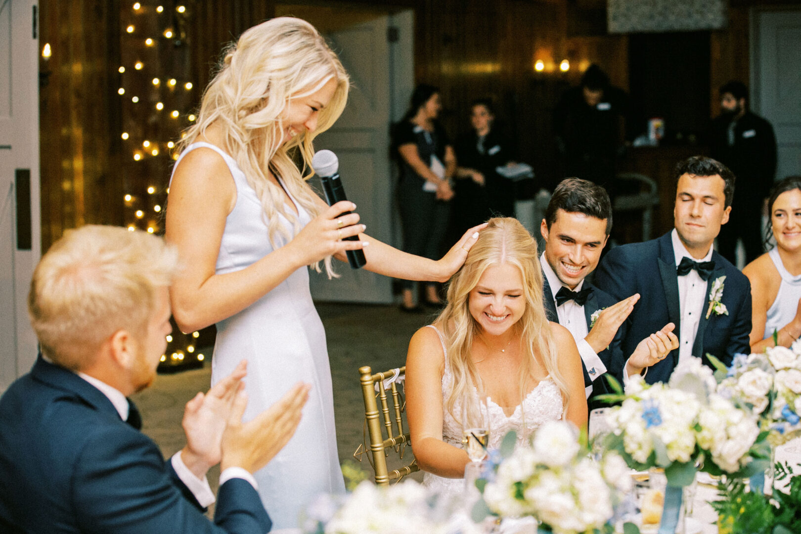 At a charming Normandy Farm wedding, a woman in a light dress speaks into a microphone, smiling warmly as she touches the seated woman's head. The decorated room is filled with applause and joy from guests celebrating the special moment.