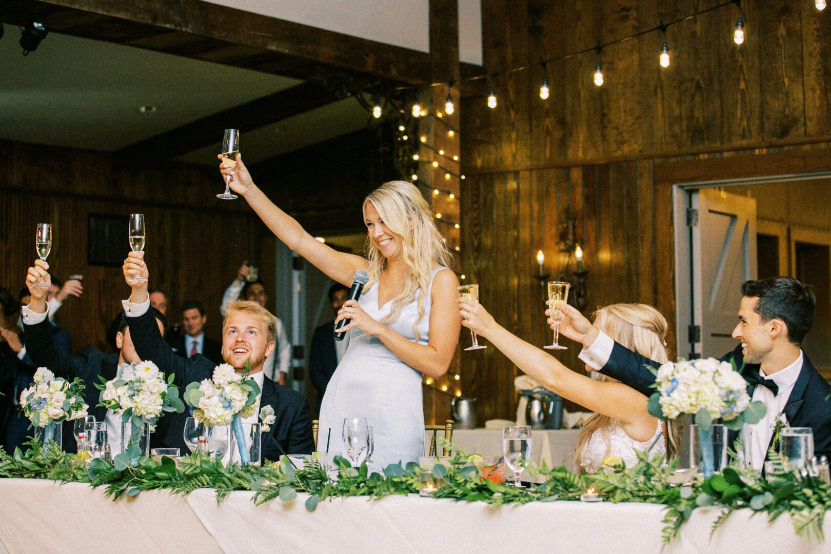 A woman in a light blue dress stands at a table, holding a microphone and raising a glass in a toast amidst the enchanting ambiance of string lights and greenery. It's the perfect setting for this elegant Normandy Farm wedding, as others seated around her join in raising their glasses.