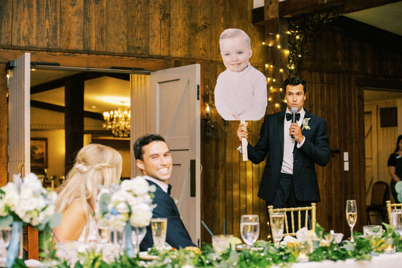 At a delightful Normandy Farm wedding, a man in a suit holds a large cutout of a baby's face while speaking at the reception. Nearby, the happy couple sits at a table adorned with floral decorations and beverages.