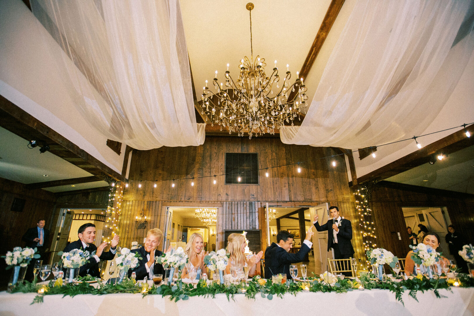 A Normandy Farm Wedding featuring a beautifully decorated table, guests seated and clapping, and a heartfelt speech being delivered under elegant chandeliers and draped fabric.