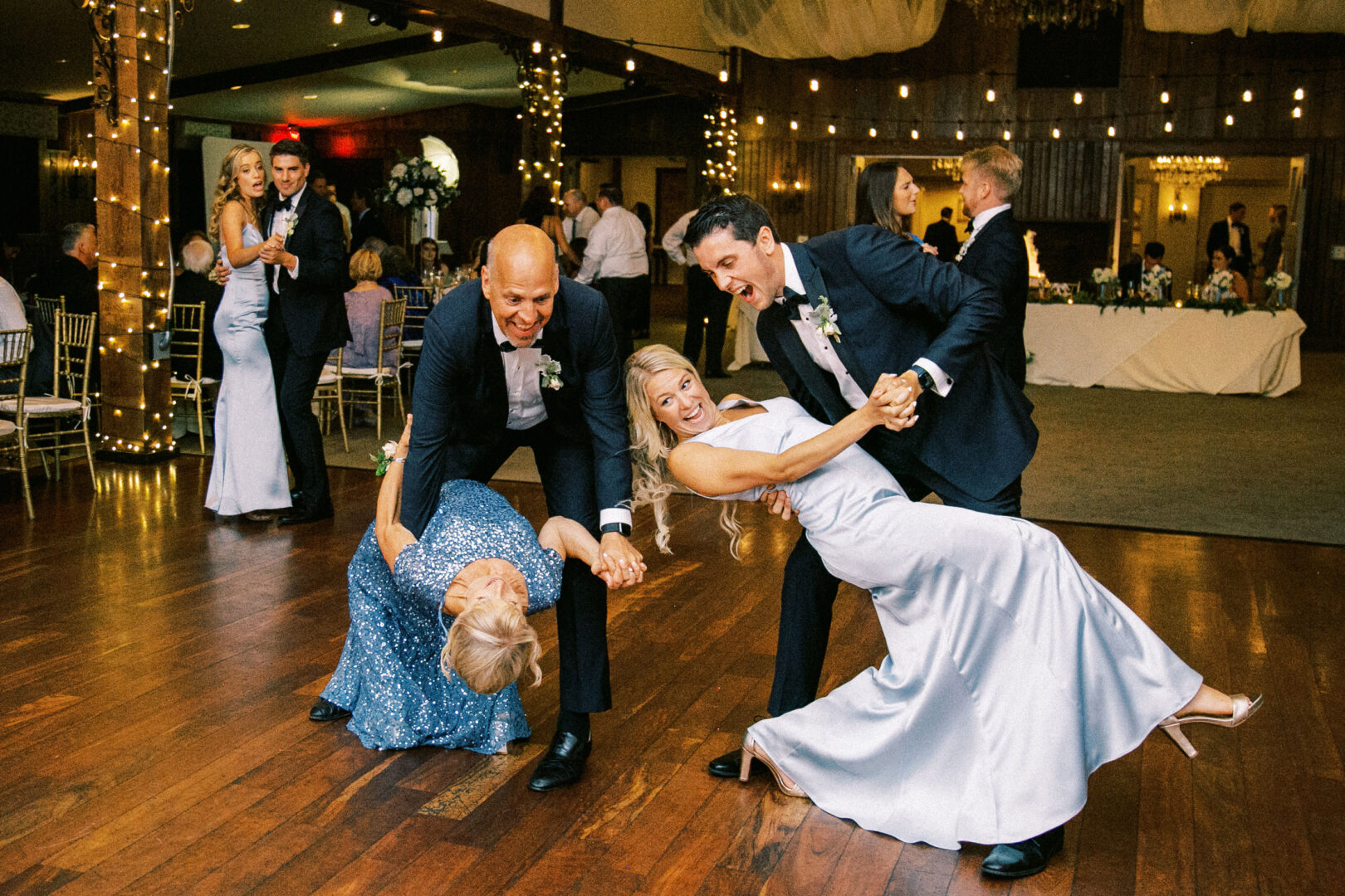 Two couples in evening attire dance enthusiastically on the wooden floor at a Normandy Farm wedding, with twinkling lights adding a magical touch to the decorated indoor event.