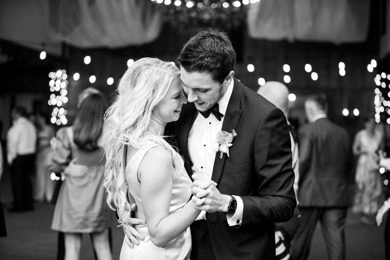 At a Normandy Farm wedding, a couple dances closely amid the gathered guests. The man wears a suit with a boutonniere, complementing the woman’s elegant sleeveless dress in the softly lit venue.