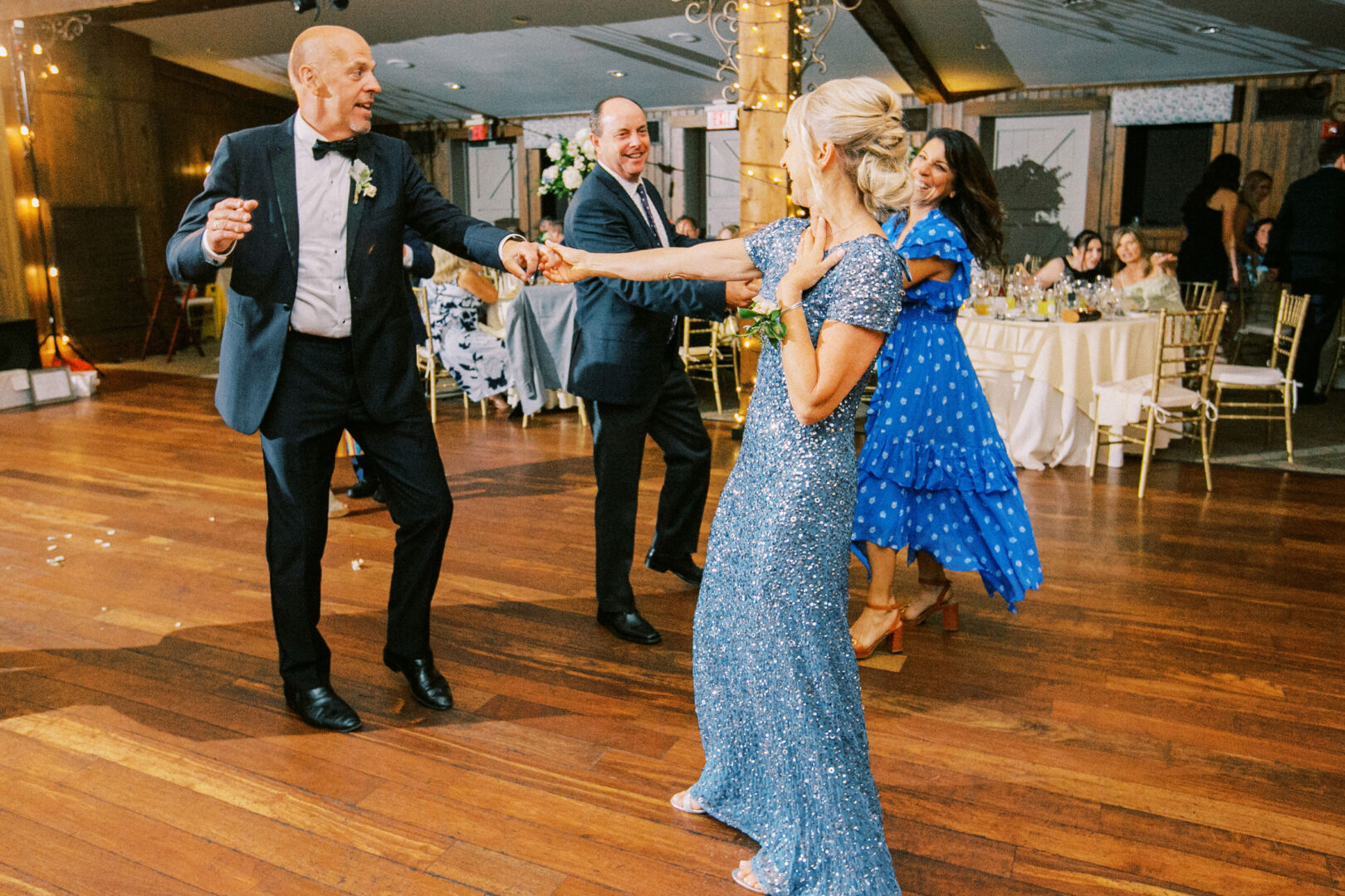 Four people in formal attire are dancing joyfully on a wooden floor at an indoor Normandy Farm wedding, with string lights and elegantly set tables in the background.